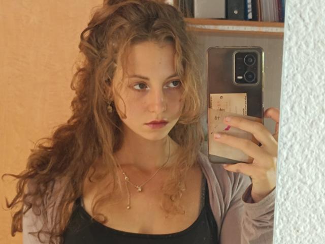 MaoLucky69 met krullend bruin haar, kijkt in de spiegel, houdt een telefoon vast, draagt een zwarte top en een roze vest, toont kleine borsten, heeft bleke huid, subtiele make-up, een serieuze uitdrukking, op een selfie..