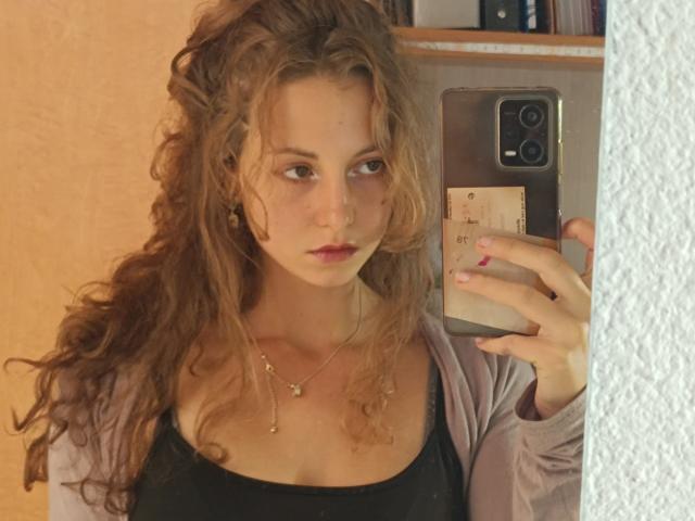 MaoLucky69 with curly brown hair, wearing a black top and purple cardigan, taking a mirror selfie with a serious expression, holding a phone with a beige case.