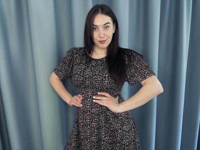 KaleyReed with dark brown hair, wearing a black leopard print dress, hands on hips, standing against blue curtains, smiling slightly, blue eyes, medium breasts, light skin.