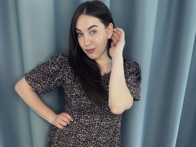 KaleyReed with long dark brown hair, wearing a leopard print dress, hand on hip, other hand touching ear, blue eyes, slight smile, standing against blue curtain.