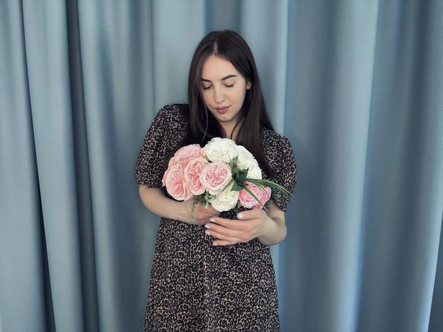 KaleyReed with long brown hair, wearing a leopard print dress, holding a pink and white rose bouquet, looking down at it, standing in front of blue curtains.