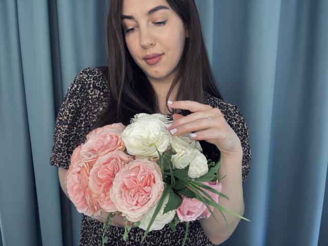 KaleyReed with long brown hair, wearing leopard print, holding pink and white roses, gently touching with right hand, blue curtain background, subtle smile, white nail polish, ring on finger.