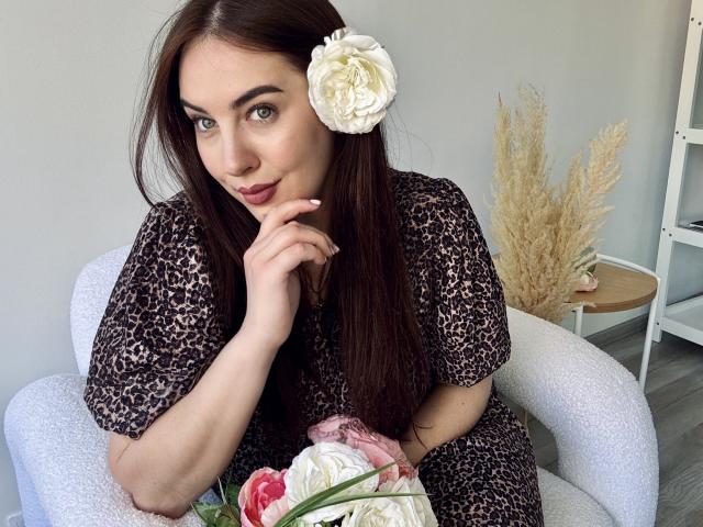 KaleyReed with brown hair, white flower in her hair, leopard print dress, thinking with finger on her lips, holding pink and white flowers, sitting on white chair.