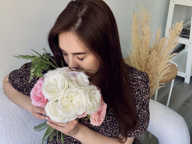 KaleyReed with long brown hair, eyes closed, holding white and pink roses, wearing a leopard print top, in a cozy indoor setting. Boobs partially visible under top.