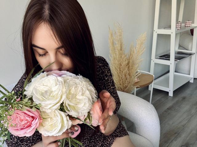KaleyReed with brown hair, wearing a leopard print dress, smelling a bouquet of white and pink roses, holding it with her right hand, in a modern room.