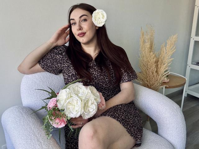 KaleyReed with long brown hair, wearing a leopard print dress, white rose hair clip, holding white and pink roses, sitting on a white chair, leg crossed.