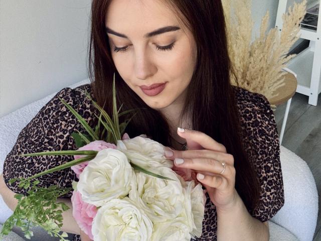 KaleyReed with dark brown hair, leopard print shirt, holding white and pink roses, slightly smiling, eyes closed, left hand with ring, pink nails, in a cozy setting.