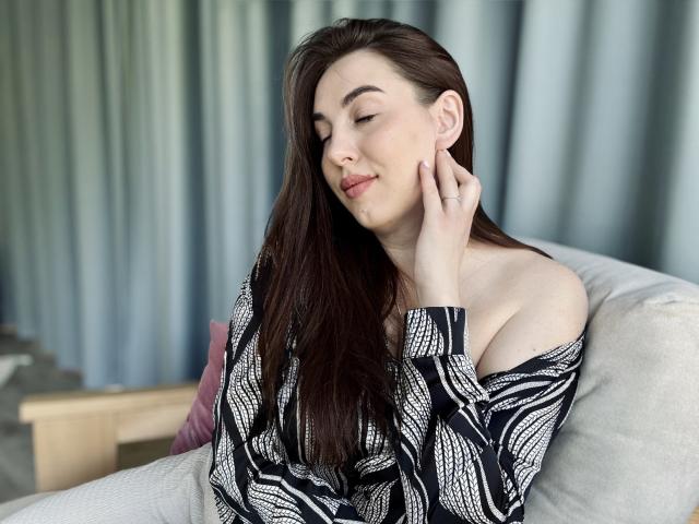 KaleyReed with long brown hair, eyes closed, smiling, off-shoulder black and white patterned top, hand touching cheek, pale skin, white couch, soft natural light, relaxed.