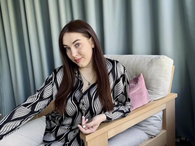 KaleyReed has long brown hair, blue eyes, and a cute smile, wearing a black and white patterned dress, sitting on a wooden chair with a pink pillow.