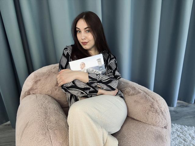 KaleyReed with long brown hair, black and white patterned blouse, white pants, sitting on a pink fluffy chair, holding a magazine, smiling, looking at camera.