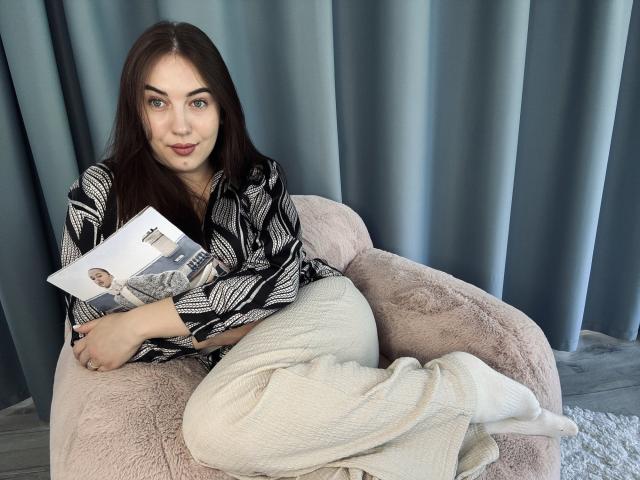 KaleyReed with brown hair, blue eyes, wearing a black and white patterned shirt and white pants, sitting in a pink furry chair, holding a magazine, showing her pussy.