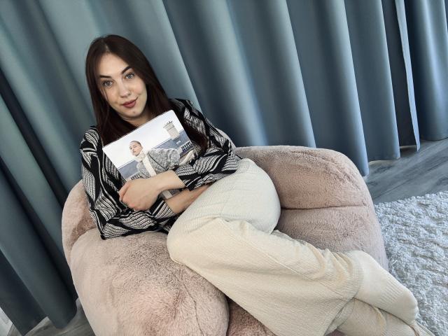 KaleyReed with long brown hair, wearing a black-and-white patterned shirt and white pants, sitting on a pink furry chair, holding a book, with a calm expression.