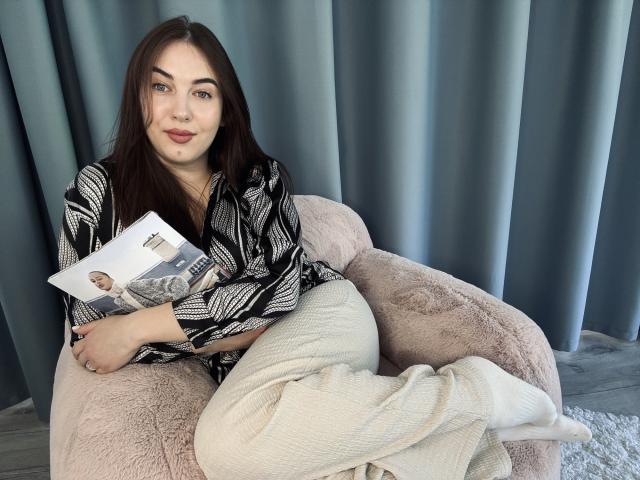 KaleyReed with dark brown hair, black and white patterned shirt, white fluffy pants, holding a magazine, sitting on a pink fuzzy chair, gray curtains in the background.