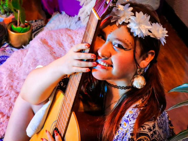 AliaMillers with red nails, white flower headband, and purple earrings, smiling while holding a guitar. She's got a black choker and is wearing a patterned dress.