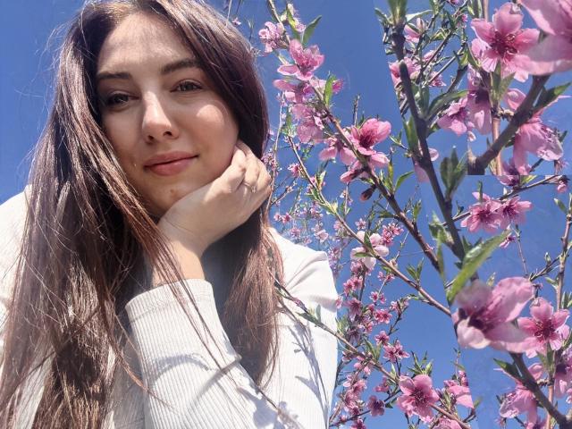 KaleyReed with long brown hair, light skin, and a slight smile, resting her chin on her hand, wearing a white sweater, next to pink cherry blossoms under a blue sky.