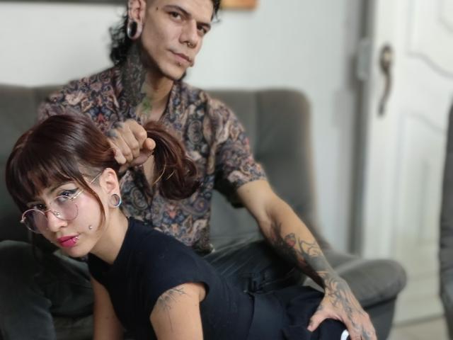 RayAndNexxy with brown hair, glasses, and pink lipstick, bent over on a gray couch, getting her hair pulled by tattooed RayAndNexxy in a patterned shirt, holding her ponytail.