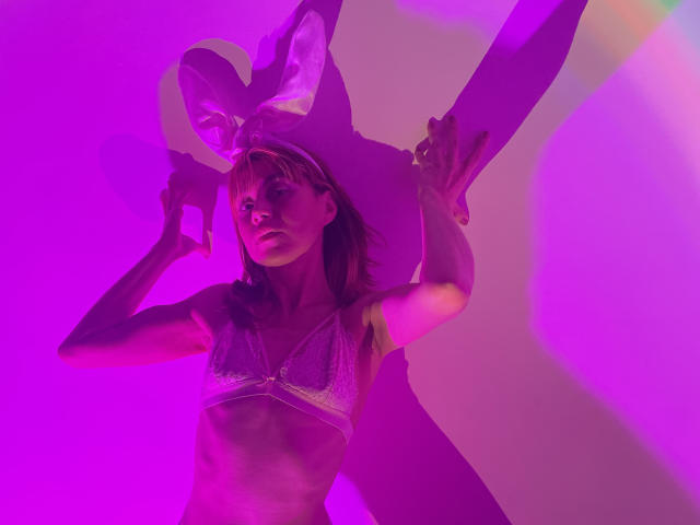 MaryAndrea with pink-lit skin, wearing a white lace bra, has her arms raised, sporting a bunny ear headband, casting a shadow of herself on the wall.