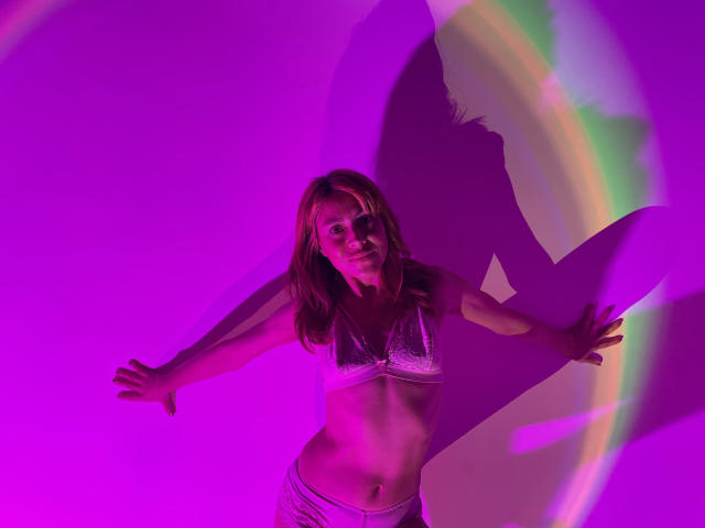 MaryAndrea with small tits, wearing white lace bra and white pants, arms out, shadow of her face and hair on purple-lit wall, looking confident and sexy.