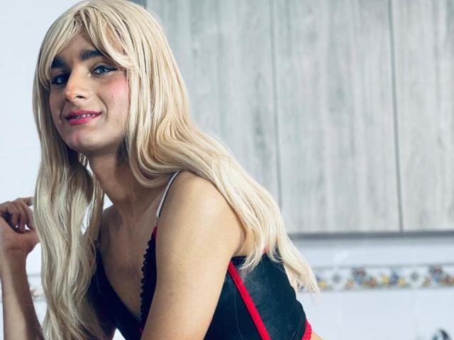 MiaMalkoba with long blonde wig, pink lipstick, black and red dress, smiling, blue eyes, light skin, small tits, in a kitchen, wooden cabinets in the background.