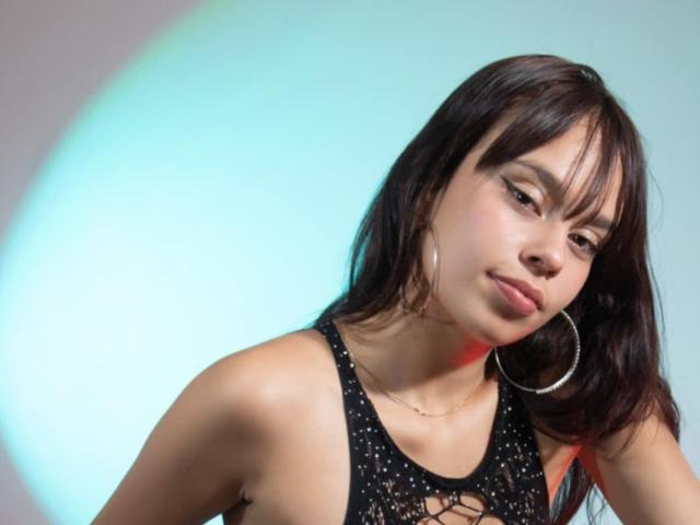 SophiaBianca with dark brown hair, small hoop earrings, and a black, sparkly top, looking slightly to the side with a subtle smile. Her ass is not visible.