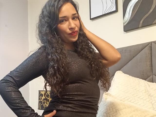Zoey Moretti, com longos cabelos cacheados e pretos, vestindo uma blusa preta brilhante de manga comprida, com a mão na cintura, sorrindo, batom vermelho, em pé em um quarto com uma cabeceira cinza..