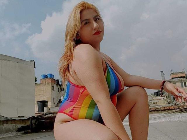 SharonMendezS with big ass, wearing a rainbow-striped one-piece, squatting, blonde hair, red lipstick, light skin, looking at camera, urban rooftop background.