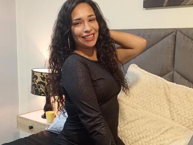 Zoey Moretti, com longos cabelos cacheados e pretos, sorrindo, vestindo uma blusa preta brilhante de manga comprida, sentada em uma cama com um travesseiro branco acolchoado, com a mão no cabelo..