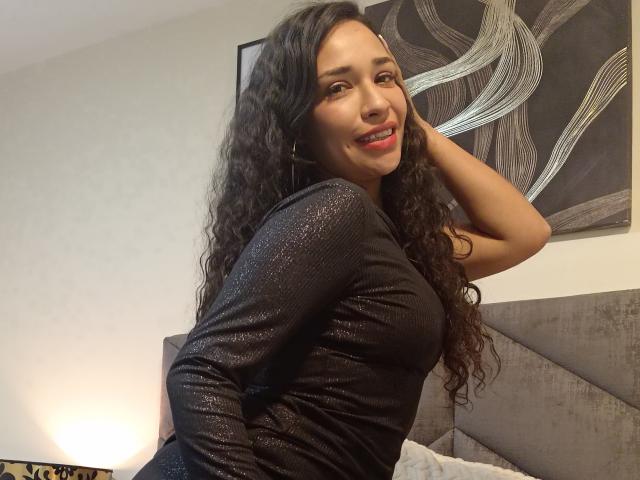Zoey Moretti mit lockigem, schwarzem Haar, lächelnd, trägt ein glitzerndes, schwarzes Oberteil mit langen Ärmeln, hat eine Hand im Haar, sitzt auf einem Bett, schaut zurück und zeigt ihr Gesäß..