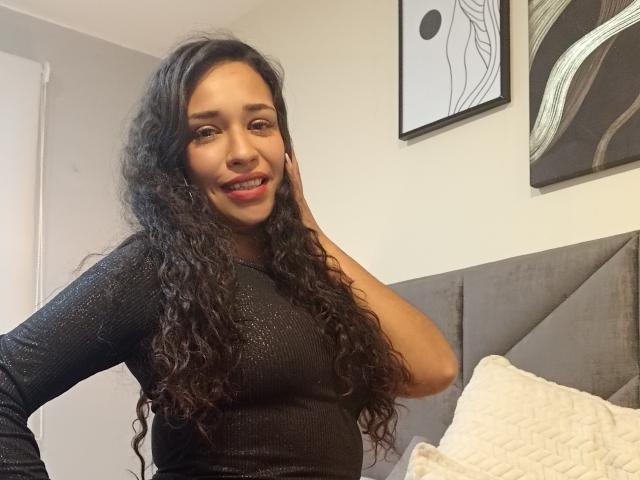 Zoey Moretti mit langen, lockigen, schwarzen Haaren, trägt ein schwarzes, glitzerndes Oberteil mit langen Ärmeln, lächelt in die Kamera, ihre Hand berührt ihr Gesicht, im Hintergrund ist ein grauer Kopfteil und ein weißes Kissen zu sehen..