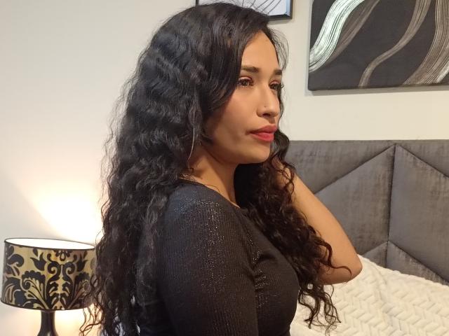 Zoey Moretti mit langem, lockigem, schwarzem Haar, die ein glitzerndes schwarzes Oberteil trägt, blickt über ihre Schulter, hat rote Lippen, weiches Licht, ein graues Kopfteil und eine schwarze, gemusterte Lampe im Hintergrund..