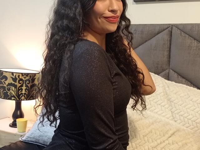 Zoey Moretti, com longos cabelos cacheados e pretos, vestindo uma blusa preta brilhante, sentada em uma cama, sorrindo, exibindo seus seios, com uma cabeceira cinza e uma luminária estampada..