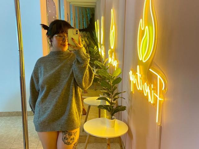 SofiLynch with glasses, short black pigtails, grey sweater, thigh tattoo, holding phone, standing in neon-lit room, small white table with plant, smiling at mirror.
