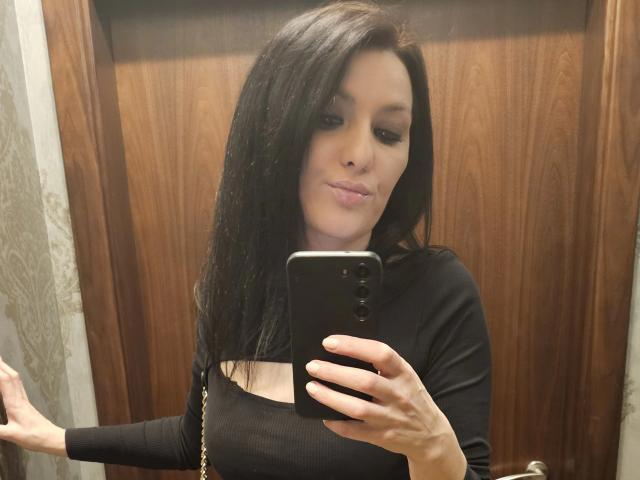 JoliDanielle with long black hair, wearing a black long-sleeve top, taking a mirror selfie with her phone, pouty lips, and a gold chain bag, showing her tits.