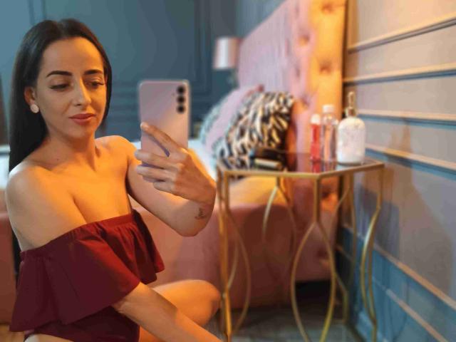 SallyMadison with dark hair, off-shoulder red ruffle top, taking a selfie, holding pink phone, sitting in pink bedroom, gold table with lotion bottles, gentle smile.