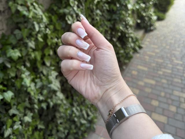 SugarrX's hand with pale pink, French-tipped nails, a small rhinstone on the ring finger, and a silver mesh bracelet, holding up to the camera.