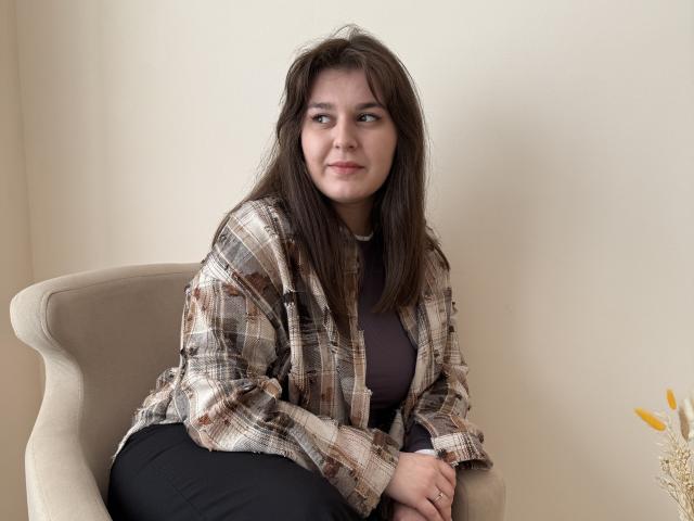 CloeBarrison with brown hair, plaid shirt, black pants, sitting on beige chair, looking to the side, slight smile, small breasts, hands on chair, soft lighting.