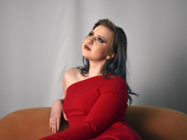 Ursula Anderson, com cabelo escuro, vestindo um vestido vermelho com um ombro descoberto, sentada em um sofá marrom, olhando para cima com uma expressão sedutora, seus seios levemente visíveis sob o vestido..