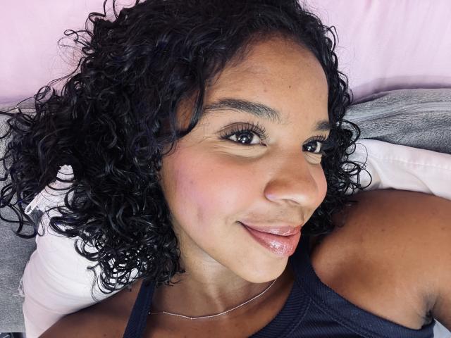 LisaMendez has curly black hair, dark skin, and a slight smile with glossy pink lips. She's wearing a black tank top and a thin necklace. Her tits are small.