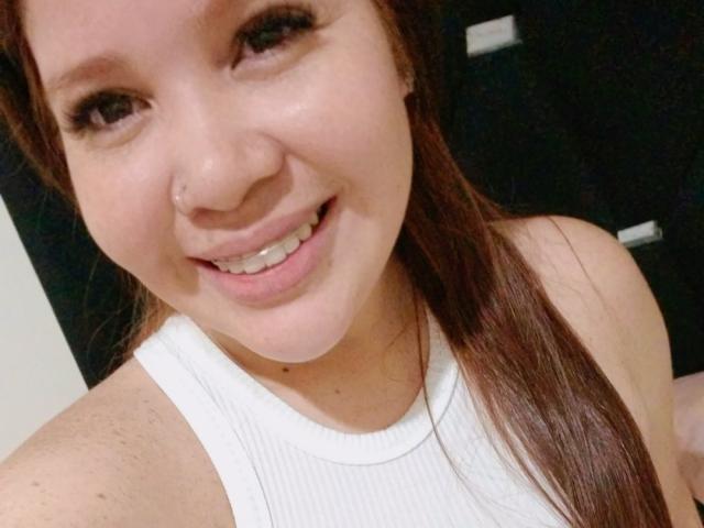 BonbonSofia with a cute smile, brown eyes, nose ring, white tank top, and long brown hair, showing her freckled shoulders and white teeth.