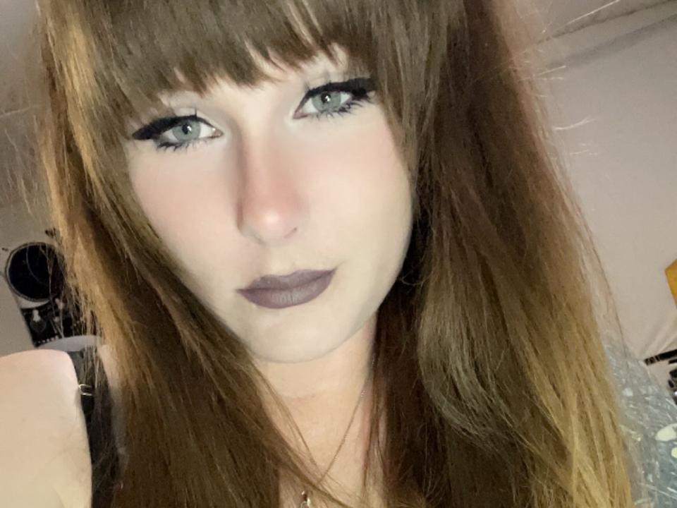 GingerrFoxy tem pele clara, olhos verdes, batom escuro e cabelo castanho com franja. Ela está olhando para a câmera com uma expressão séria, usando delineador preto..