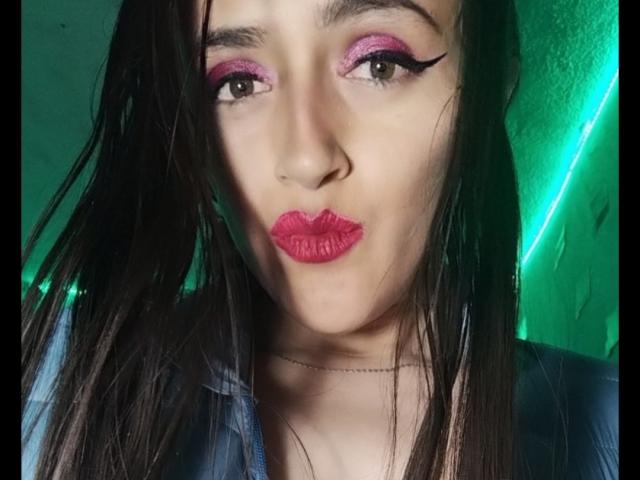 AndreaHotLove69 has pink eyeshadow, black winged eyeliner, pink lipstick, and wet black hair, wearing a blue shirt, with a green neon light in the background.