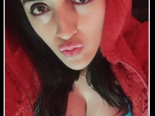 AndreaHotLove69 with pouty pink lips, brown eyes, and black hair, wearing a red fluffy hoodie, exposing cleavage, looking into the camera with a sexy, confident expression.