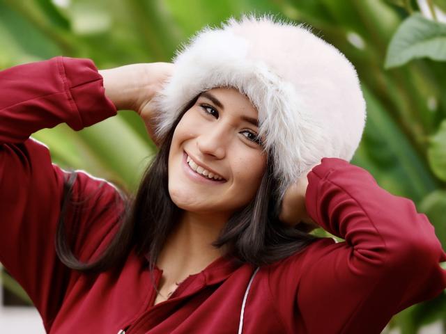 PeytonGrayy mit dunklem Haar, lächelnd, trägt einen pinken, flauschigen Hut und ein rotes Hemd, die Hände sind im Hut versteckt, zeigt ein süßes, attraktives Gesicht, eine pinke Nase und einen glücklichen Gesichtsausdruck..