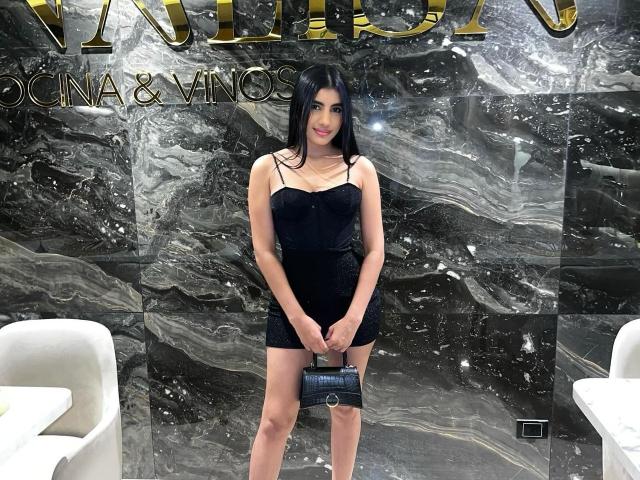 WeAreSwet: Eine Person mit langen, schwarzen Haaren, die ein schwarzes Minikleid trägt, eine schwarze Handtasche hält und vor einer Marmorwand steht, lächelnd. Brüste, Hintern und Vulva sind nicht sichtbar..