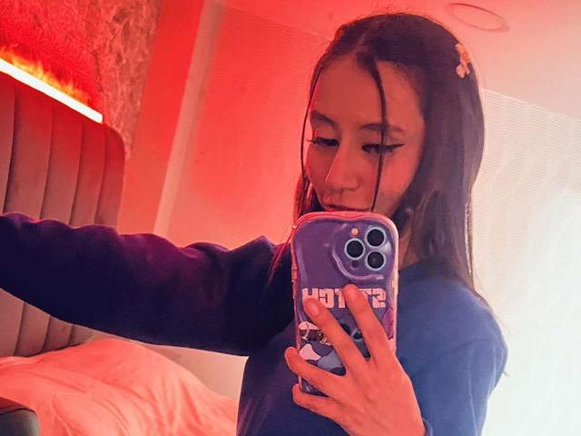 EviieGothel with long black hair, wearing a blue shirt, taking a selfie in a red-lit room, holding a purple phone with white patterns, showing off her cute face.