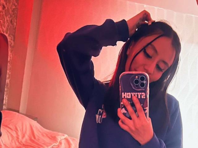 EviieGothel in a blue sweater, holding a phone with "Hoties" cover, raising one hand to her head, smiling, with long dark hair and a cute face.