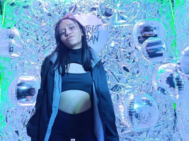 EviieGothel with long black hair, wearing a black cut-out top, showing her midriff, and a black jacket, standing in front of shiny, reflective, green-tinged walls.