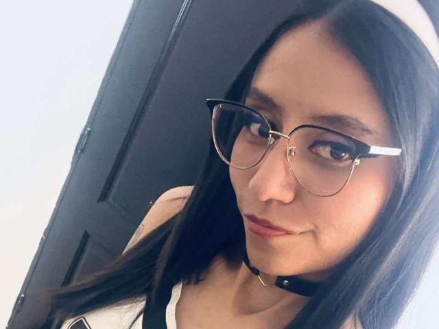 EviieGothel with long black hair, glasses, white headband, black choker, and red lipstick, looking at the camera, wearing a white tank top, showing her small tits.