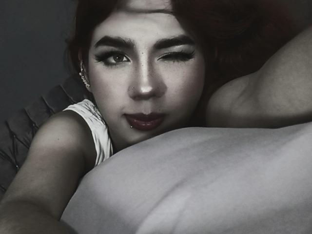 Ashleypiinnk with red hair, winking, has dark eyeliner, nose piercing, and lip ring, lying on a pillow, wearing a white sleeve, her arm and pillow in the foreground.
