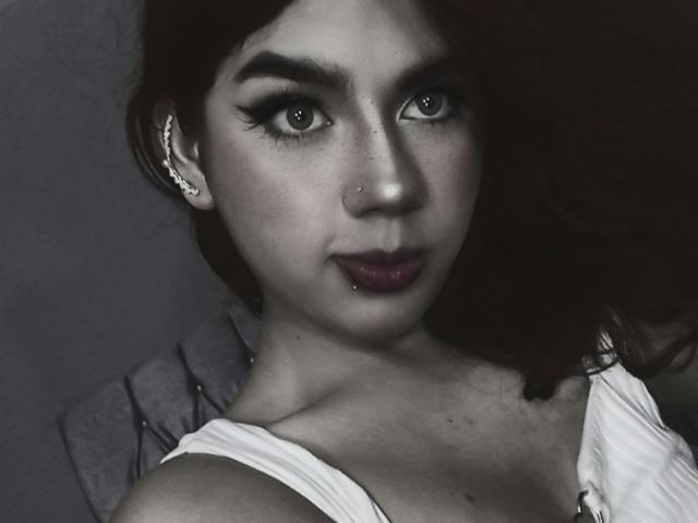 Ashleypiinnk has pale skin, red hair, dark makeup, and a nose ring. She's wearing a white top, showing her tits slightly. Her eyes are piercing, and she's looking at the camera.